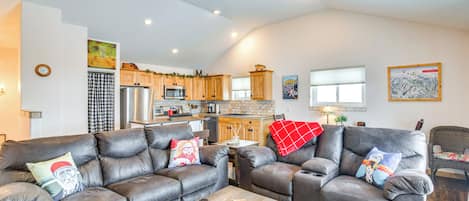 A spacious living area with two leather sofas, a wooden coffee table, and a ceiling fan. A kitchen with wooden cabinets and a refrigerator is visible in the background.