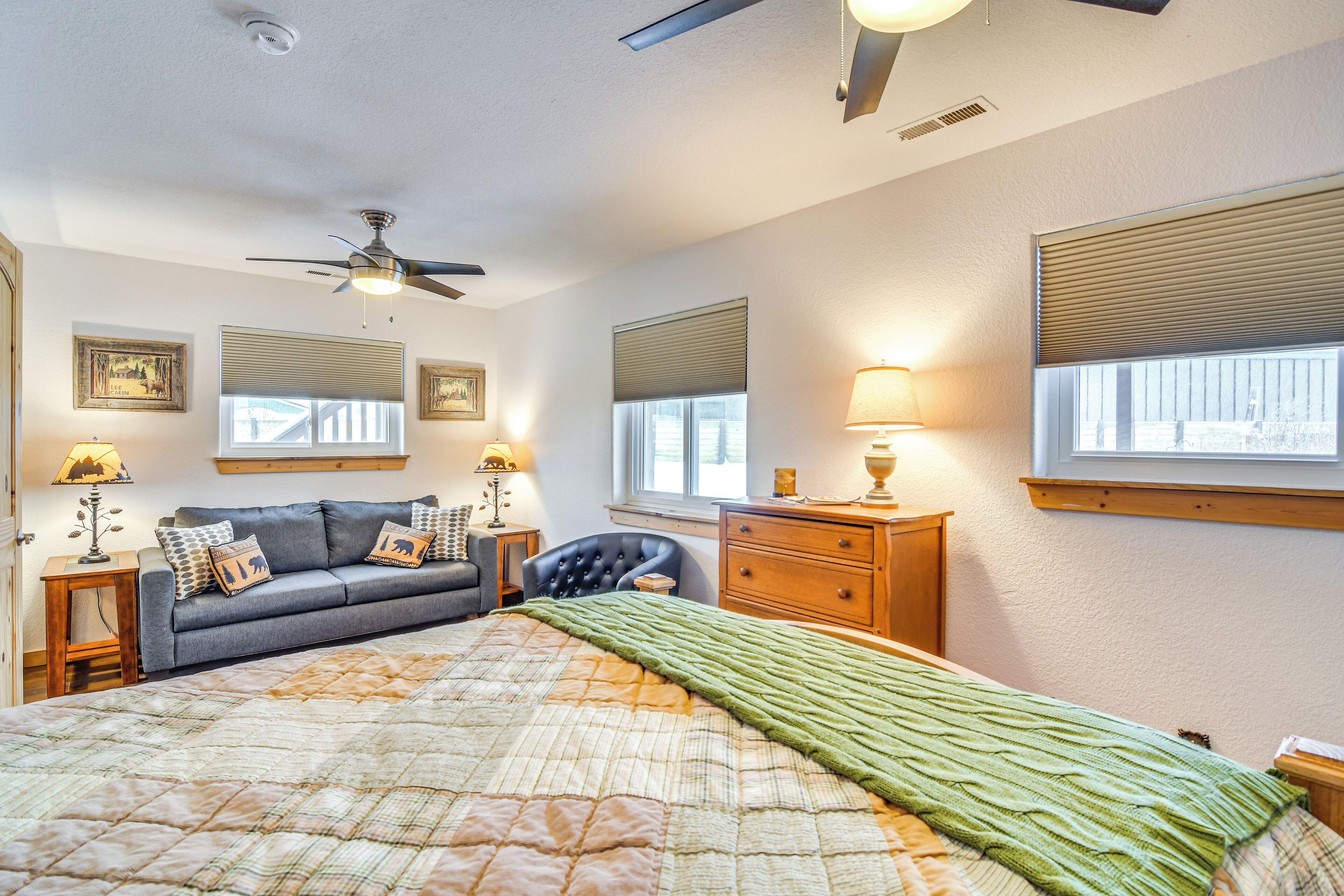 Un dormitorio con cama, sofá, cómoda, ventilador de techo y dos ventanas con persianas.