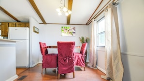 Une pièce avec un plancher en bois, un réfrigérateur blanc, un plafond en bois avec des poutres apparentes, une chaise rouge, une fenêtre avec un rideau beige et un tableau accroché au mur.