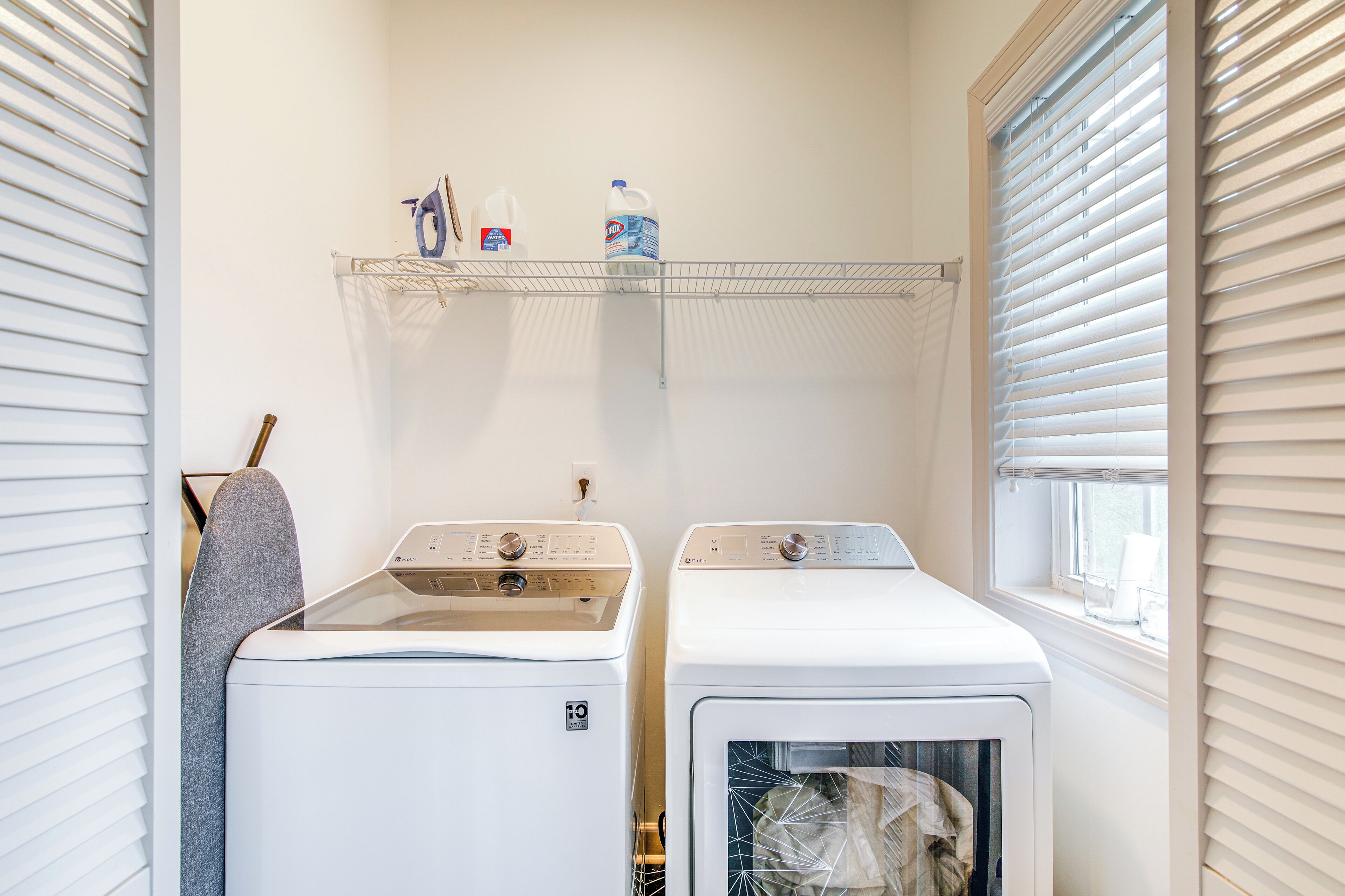 A laundry room with two white front-loading washing machines, a shelf above with cleaning supplies, and a window with blinds.