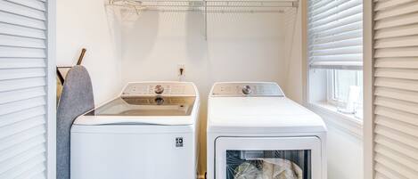 A laundry room with two white front-loading washing machines, a shelf above with cleaning supplies, and a window with blinds.