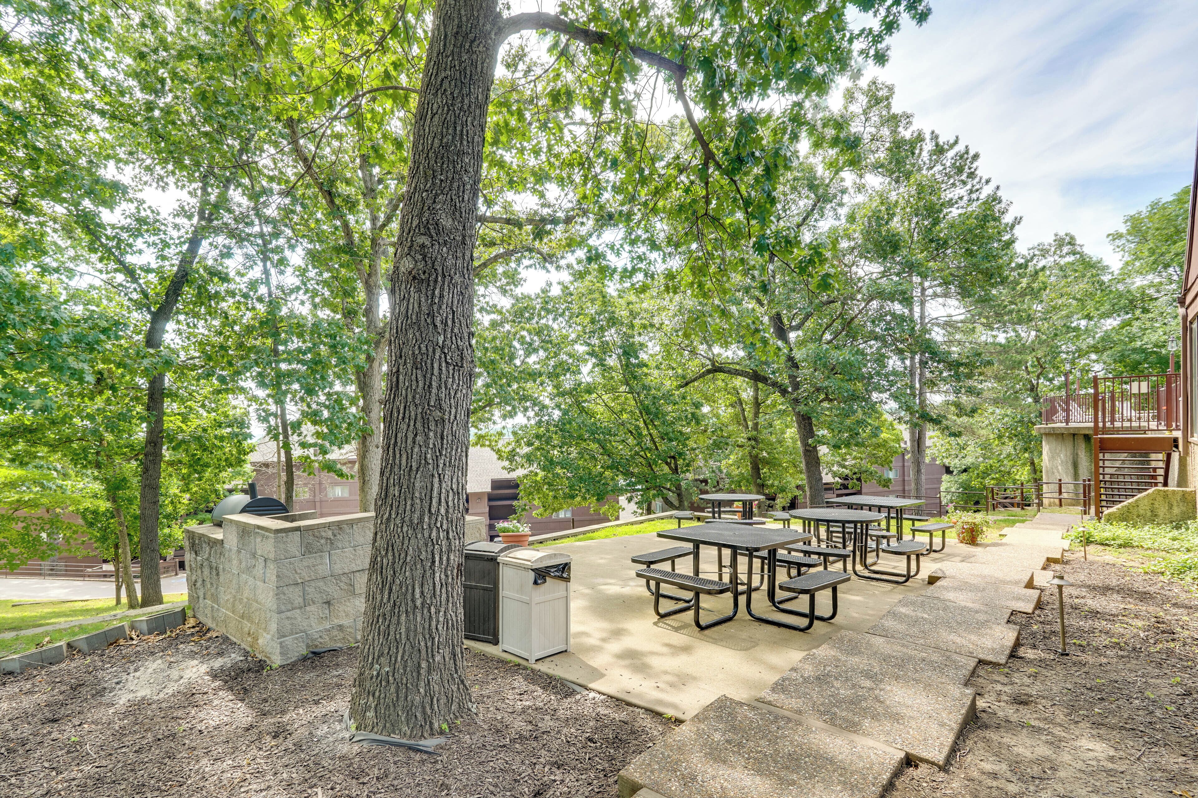 Un espace extérieur aménagé en terrasse, avec des tables de pique-nique et un barbecue, le tout entouré d’arbres.