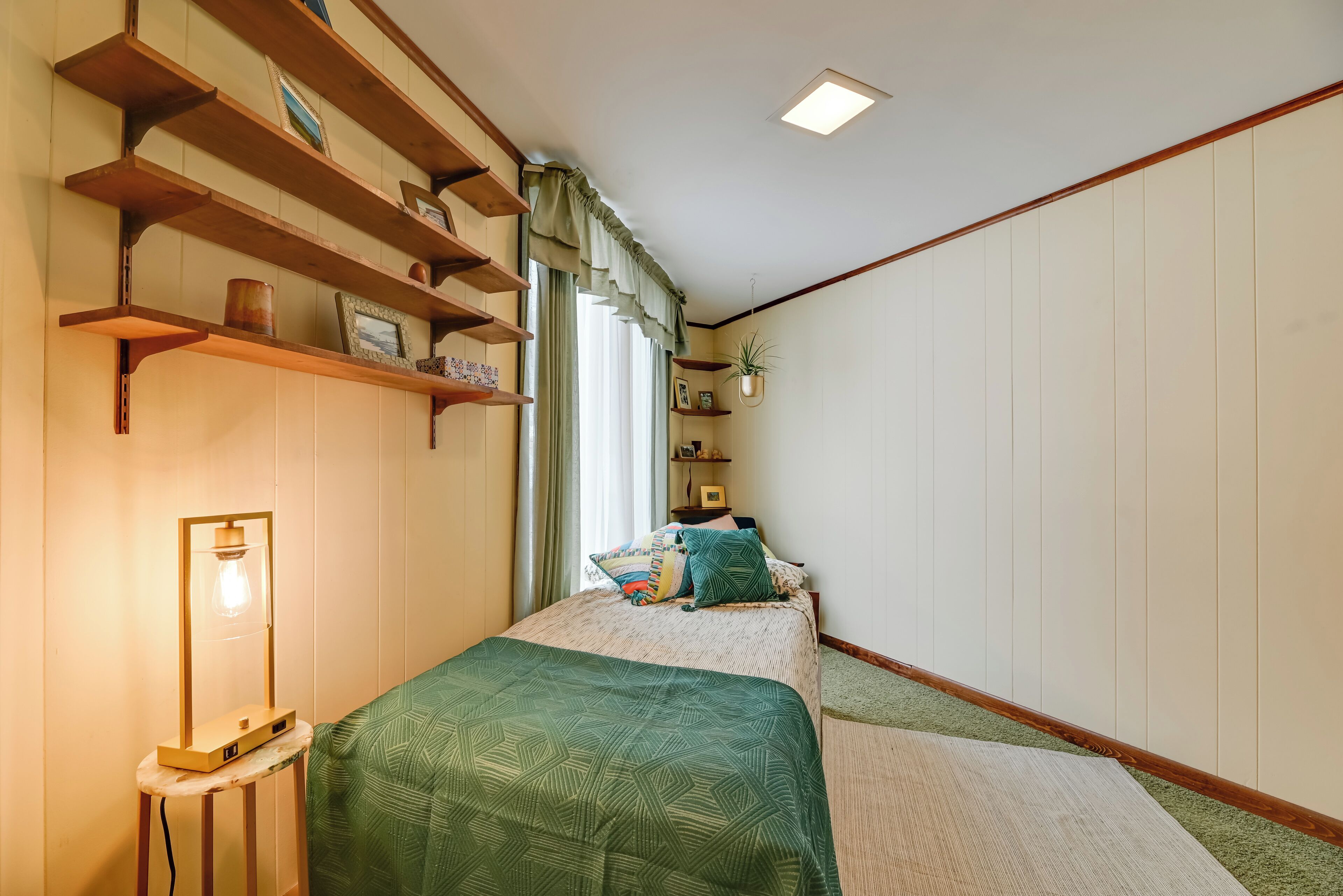 A bedroom with a bed, a bedside table with a lamp, wooden shelves, and a hanging plant.