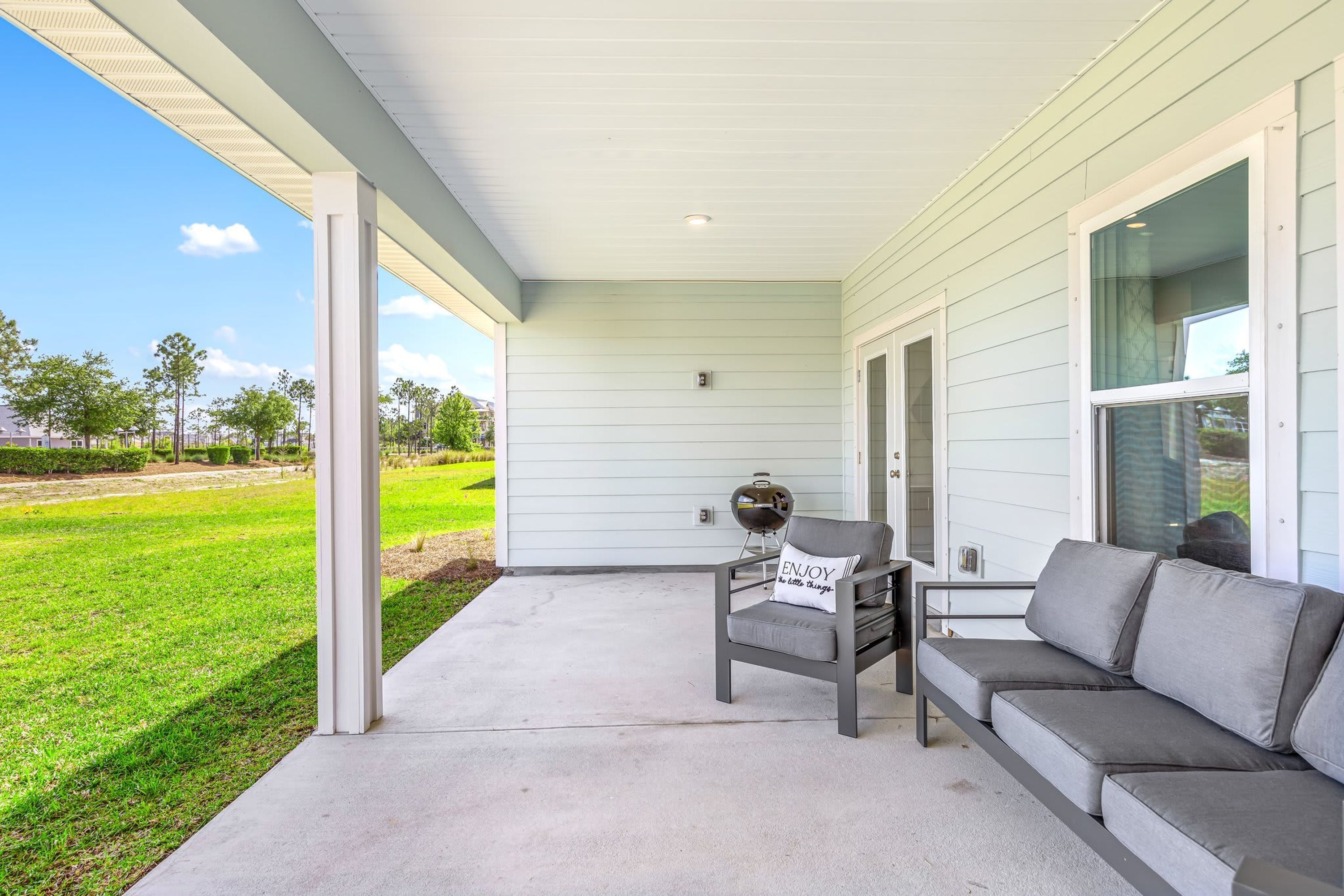 A covered porch with a gray sofa and a chair, a small table, and a window with a view of a green lawn and trees.