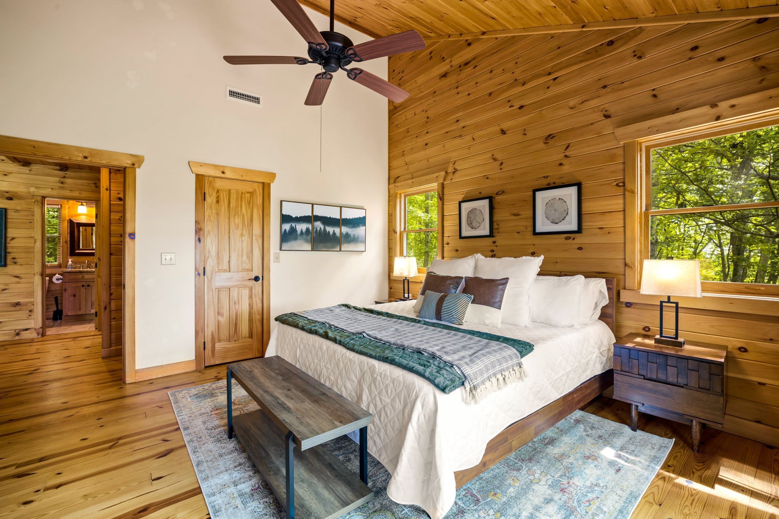 A wooden cabin bedroom with a bed, a bench, a ceiling fan, and a large window.