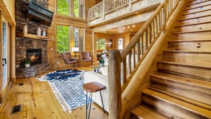 A log cabin interior with a stone fireplace, wooden staircase, and large windows. - Eagle Nest Lodge Hot Tubs Mountain Views (Banner Elk)