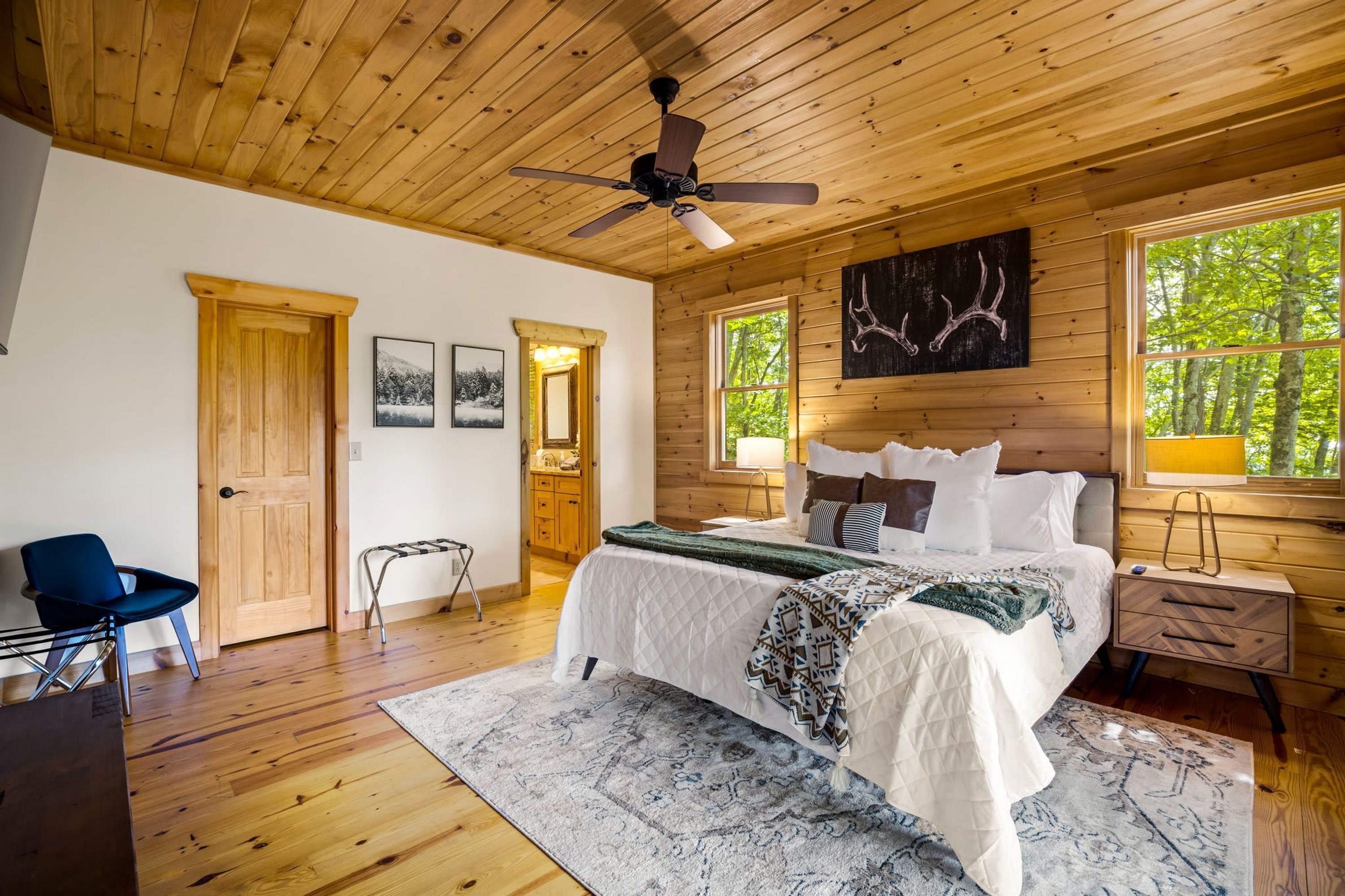 Un dormitorio tipo cabaña de madera con cama, escritorio, silla, ventilador de techo y ventana con vistas al bosque.
