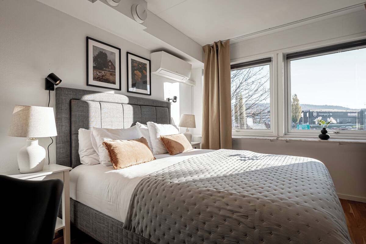 A neatly arranged hotel room with a large bed, a bedside table with a lamp, a chair, and a window with a view of a bridge.