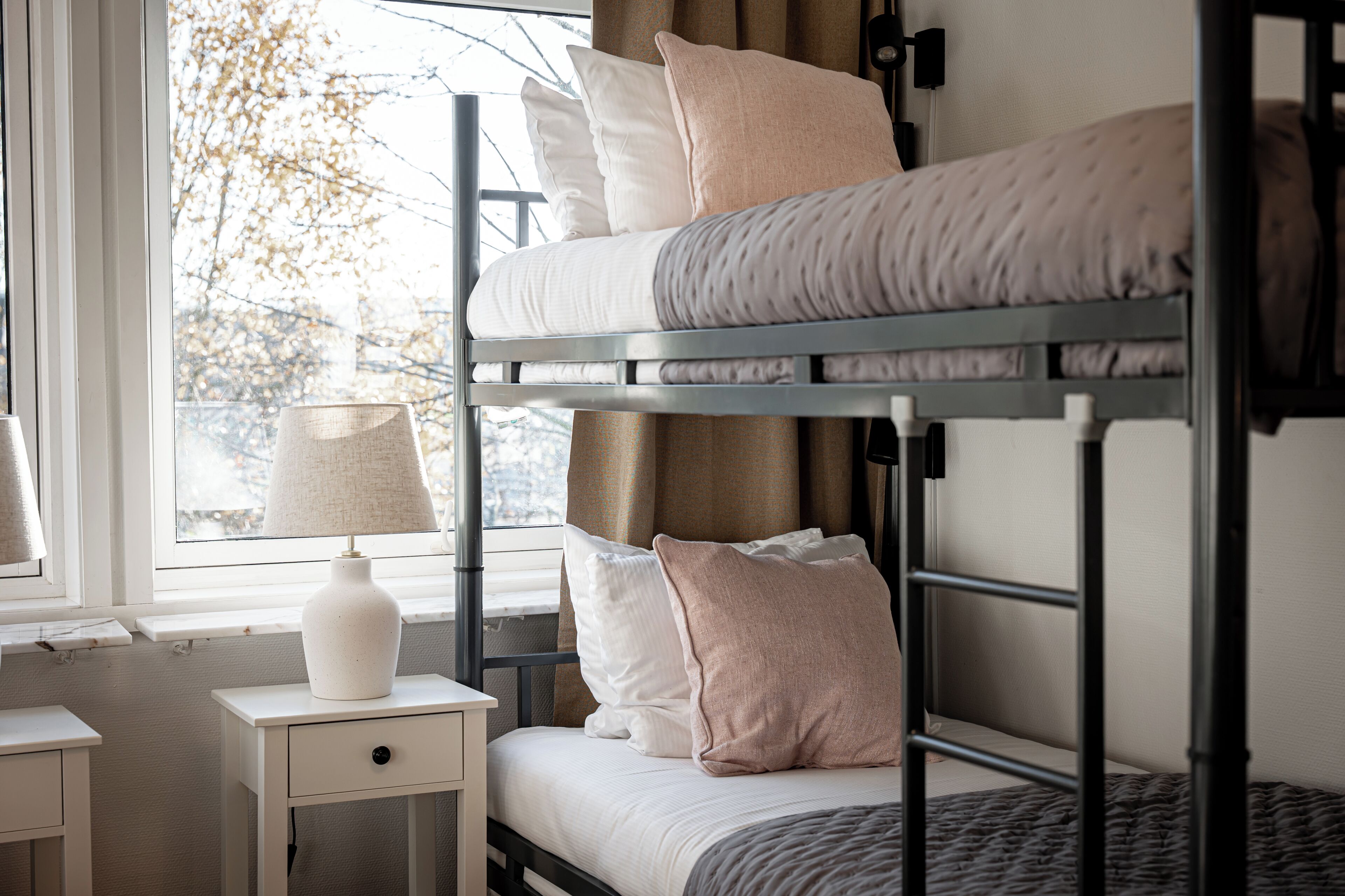 Una cama individual con almohadas, una mesita de noche con lámpara y una ventana con vista a árboles.