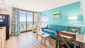 A living room with a sofa, a coffee table, and a dining table. There is a view of the ocean through large windows.