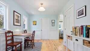 A dining area with a wooden table and chairs, a patterned rug, and a bookshelf with books. - Wellfleet 2BR Retreat – Walk to Bay & Kayak! RPMC007 (Wellfleet)