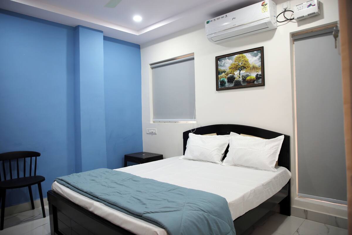 A bedroom with a bed, a chair, a small table, an air conditioning unit, and a window with blinds.