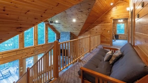 Interno di una baita con una scala in legno, un divano e un camino in pietra.
