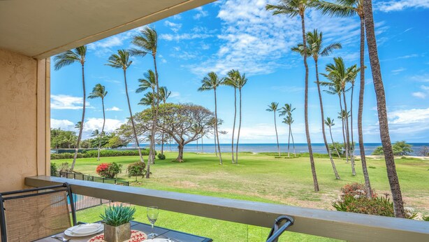 A balcony with a table set for two, overlooking a grassy area with palm trees and a view of the ocean. - Leinaala Oceanfront #205 Oceanfront Views! A/C, wifi, Pool, Great Rates! (Kihei)