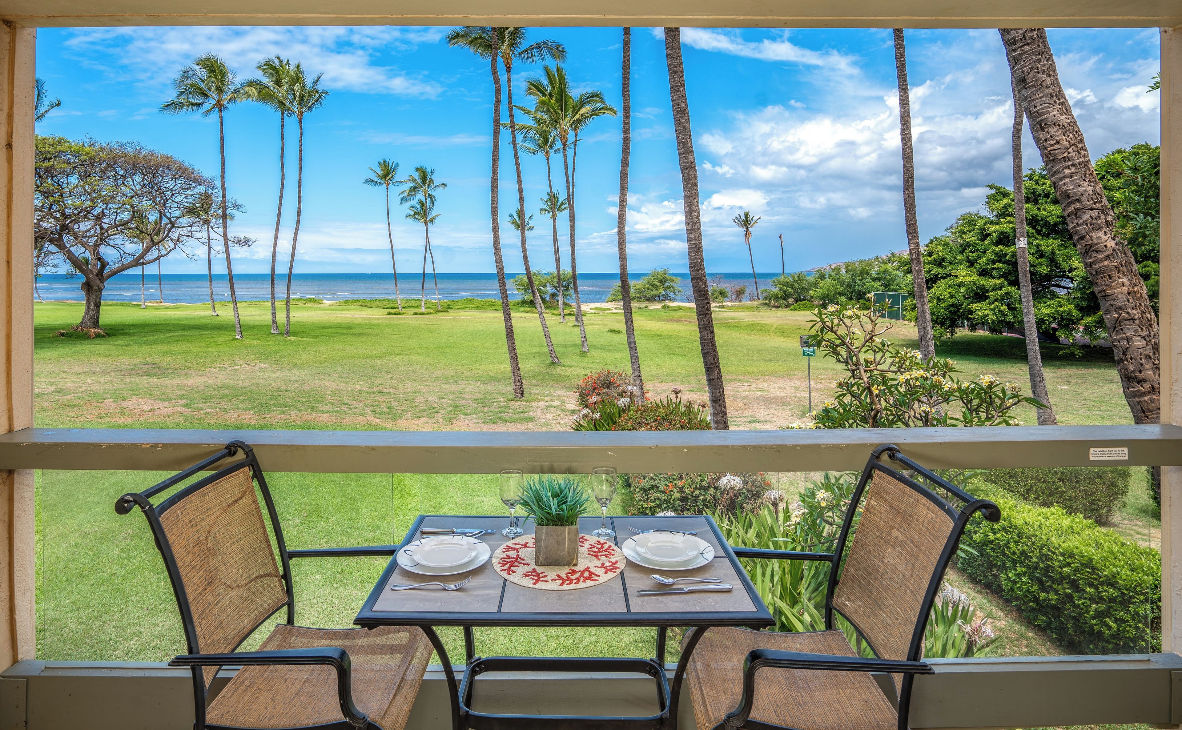 A balcony with a table set for two, overlooking a grassy area with palm trees and a view of the ocean.