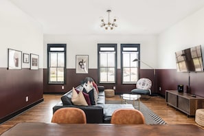 Una sala de estar moderna con piso de madera oscura, un sofá de cuero negro con muchos cojines, una mesa de centro redonda y un televisor montado en la pared.