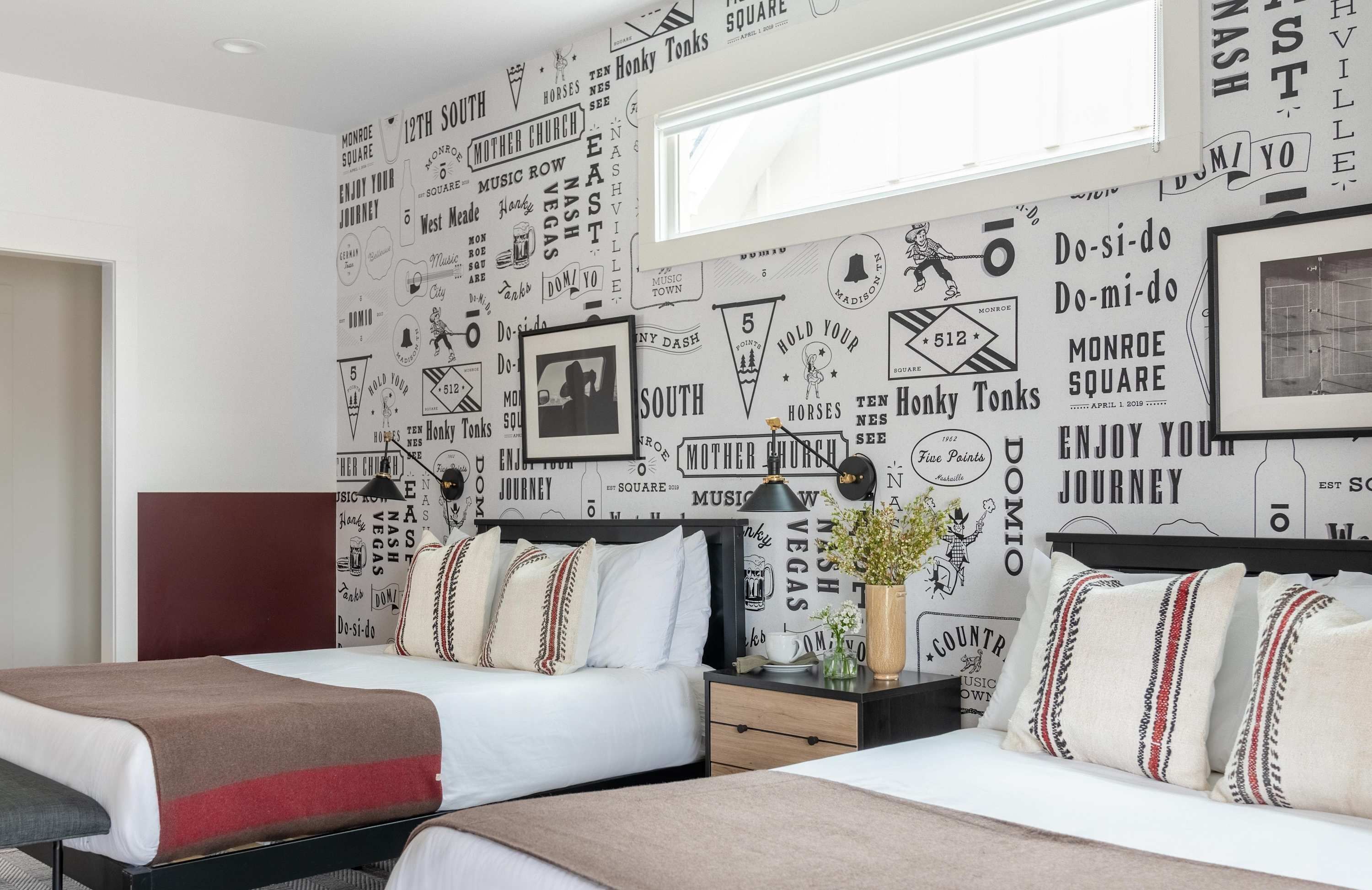 A hotel room with two beds, a nightstand, a lamp, and a wall decorated with various travel-related phrases and images.