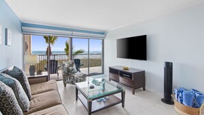 Salón con sofá, mesa de centro y televisor, con vistas a una playa con palmeras.