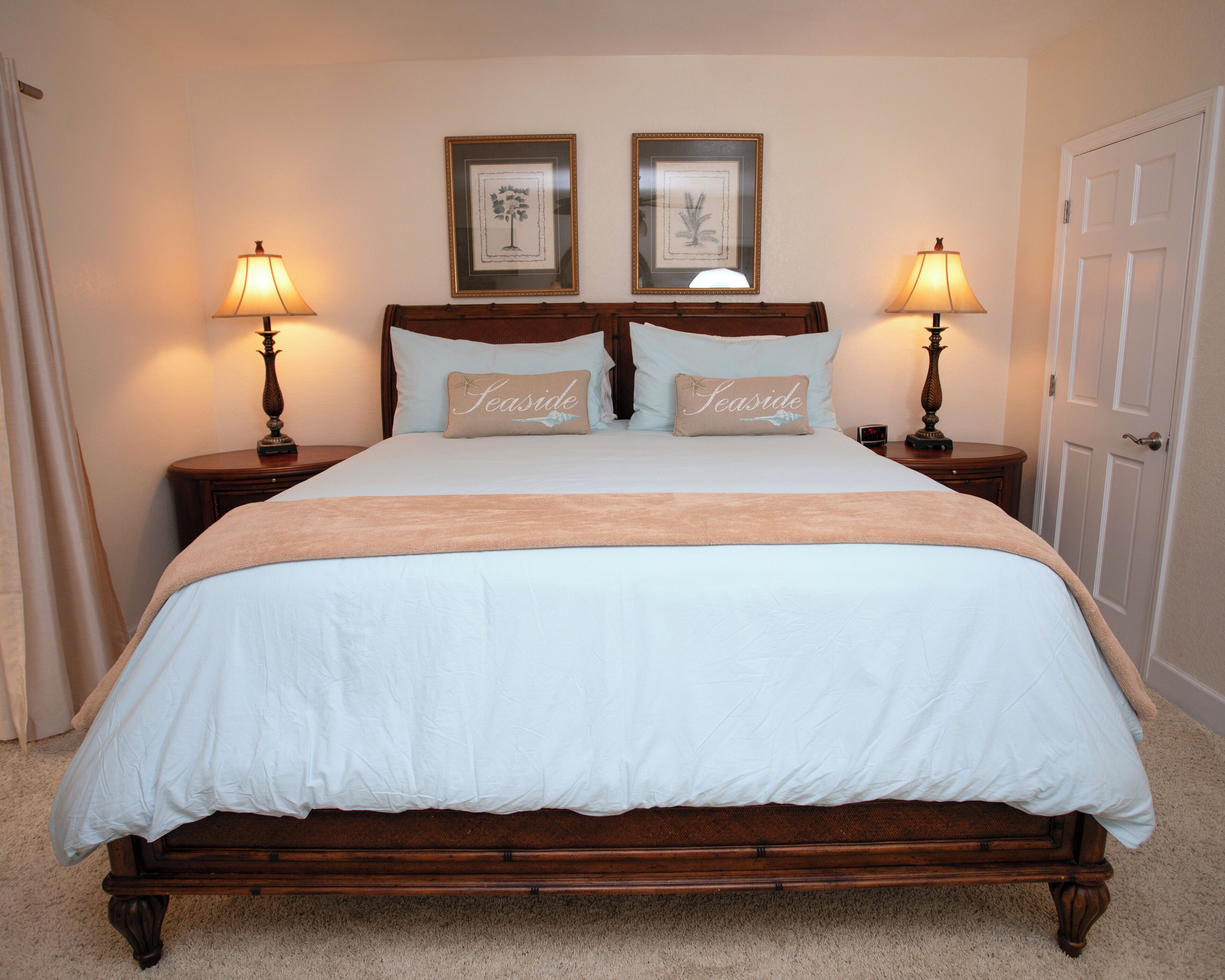 A neatly made bed with a white duvet and two pillows labeled "Seaside." There are two framed pictures on the wall above the bed, a wooden nightstand with a lamp on each side, and a door to the right.