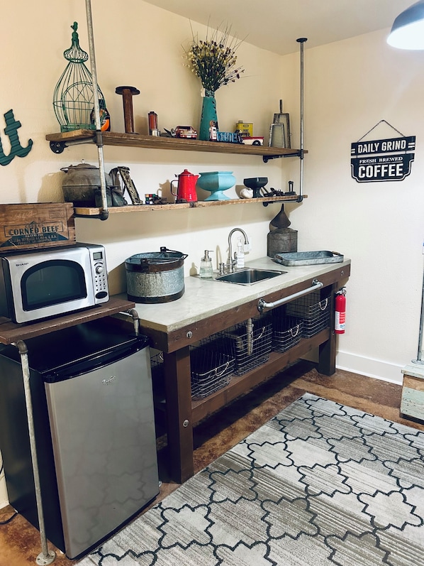 A kitchen with a microwave, refrigerator, sink, and various kitchen items on shelves. - "Milam Mill Haus” : 1925 Historic German home “Large Hot Tub”. (Fredericksburg)
