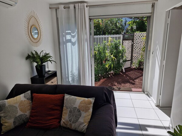 A living room with a sofa, a potted plant, and a view of a garden through a sliding door. - Luxury apartment 25 meters from the island's most beautiful beach, Boucan Canot. (Saint-Paul)