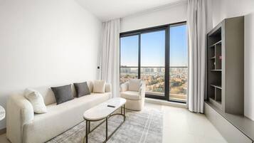 Un salon moderne comprenant un canapé, un fauteuil et une table basse, de grandes fenêtres offrant une vue sur la ville et une étagère intégrée pour la télévision.