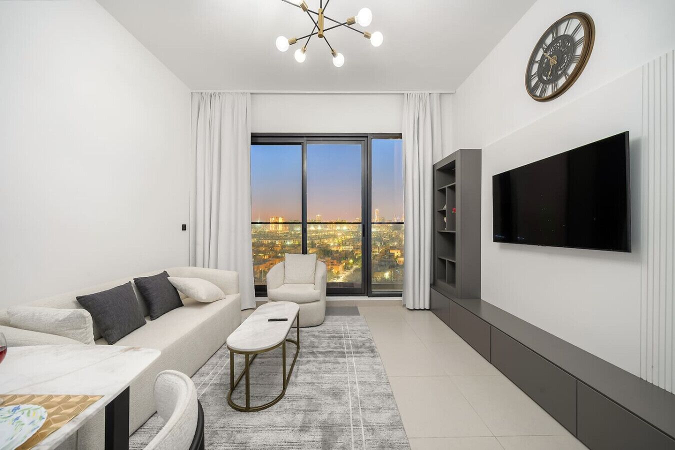 Un salon moderne avec un canapé, un fauteuil et une table basse, un téléviseur à écran plat et une vue sur la ville grâce à de grandes fenêtres.