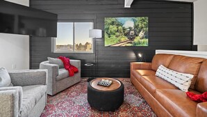A modern living room with a grey sofa, a brown leather couch, and a chess table. - Fireside Haven | Hot Tub + Downtown Walkable (Custer)