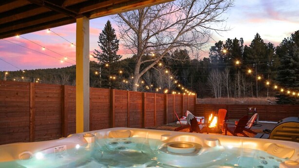 A hot tub with a wooden deck and string lights, surrounded by a wooden fence and trees. - Fireside Haven | Hot Tub + Downtown Walkable (Custer)