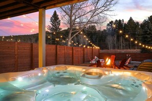 A hot tub with a wooden deck and string lights, surrounded by a wooden fence and trees.