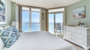 A bedroom with a large bed, a dresser, a cross on the wall, and a view of the ocean through the sliding glass door. - Baywatch Resort #1705 3 Bedroom Condo (North Myrtle Beach)