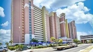 A multi-story hotel with a modern architectural design, featuring a prominent entrance and a parking area in front. - Baywatch Resort #1705 3 Bedroom Condo (North Myrtle Beach)