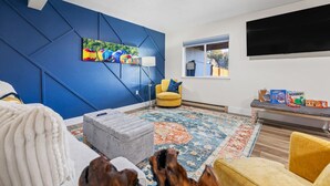 Un salon moderne avec un mur d’accent bleu, un grand téléviseur à écran plat, un fauteuil jaune et un tapis coloré.