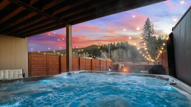 Un jacuzzi bajo una pérgola de madera con vistas al atardecer, rodeado por una valla de madera y luces cálidas.