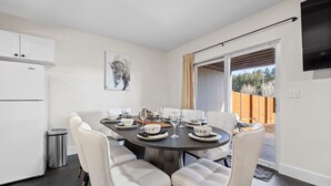 Un comedor con una mesa redonda dispuesta para cuatro personas, sillas blancas y una cocina con nevera y armarios blancos.