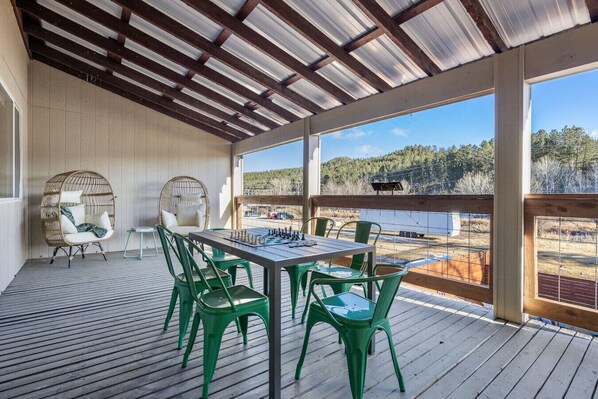 A covered outdoor deck with a chess table and chairs, overlooking a scenic landscape.