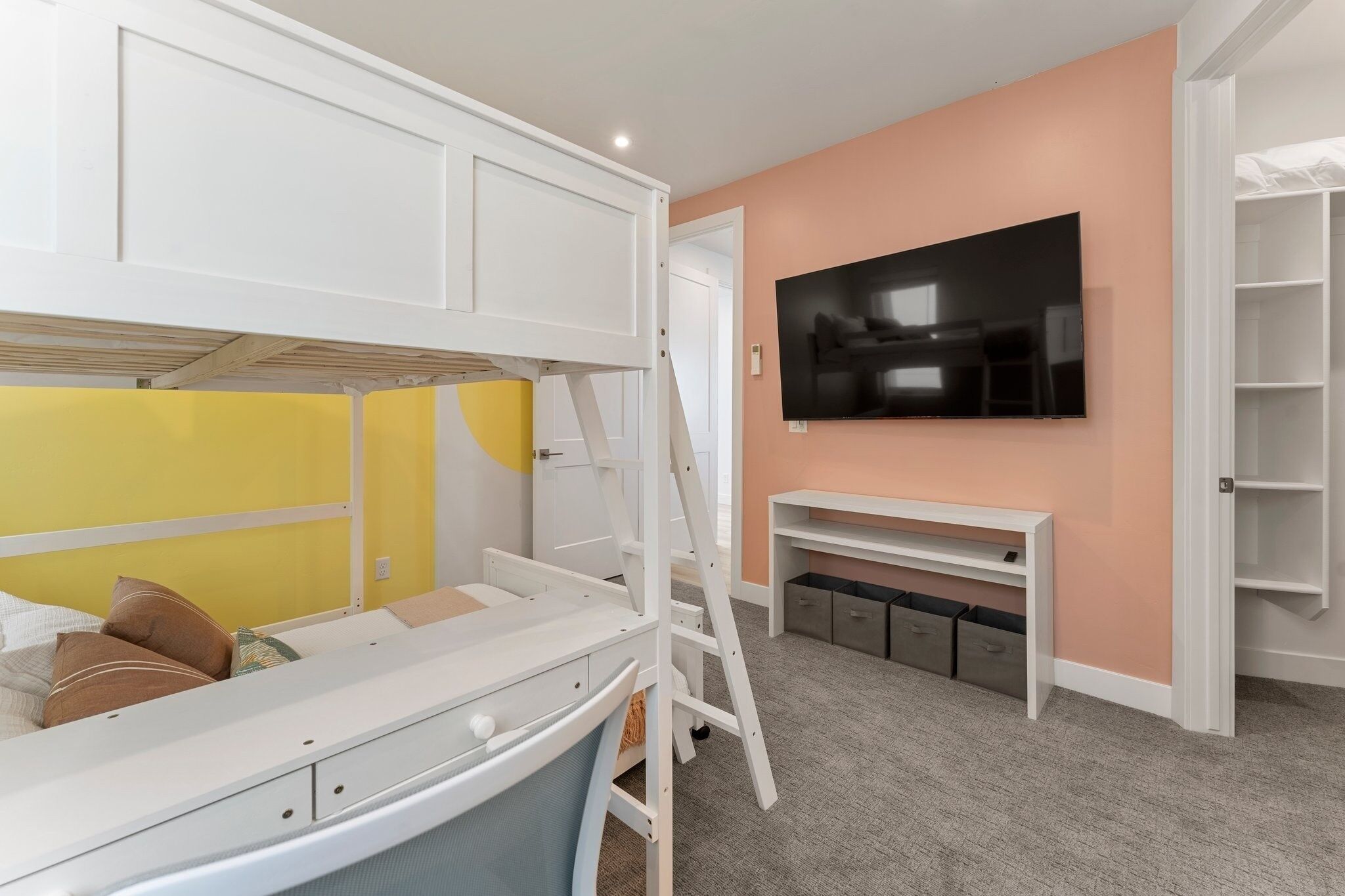 Un lit superposé pour enfant avec un bureau et une échelle, un téléviseur fixé au mur et un meuble de rangement avec des étagères.