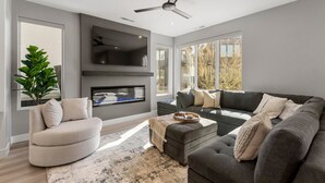 Een moderne woonkamer met een grijze bank, een beige fauteuil en een flatscreen televisie aan de muur.