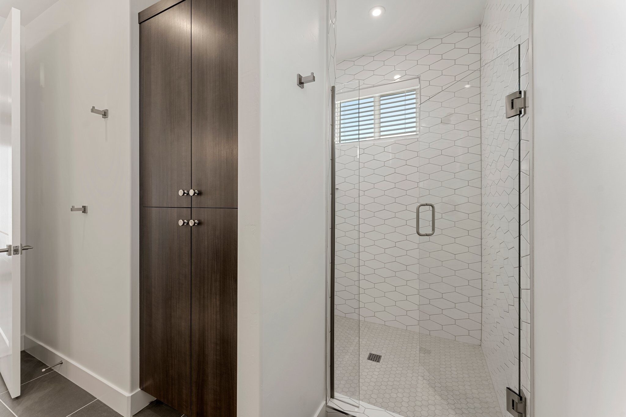 A modern bathroom with a glass shower enclosure, a wooden wardrobe, and a hexagonal tile wall.
