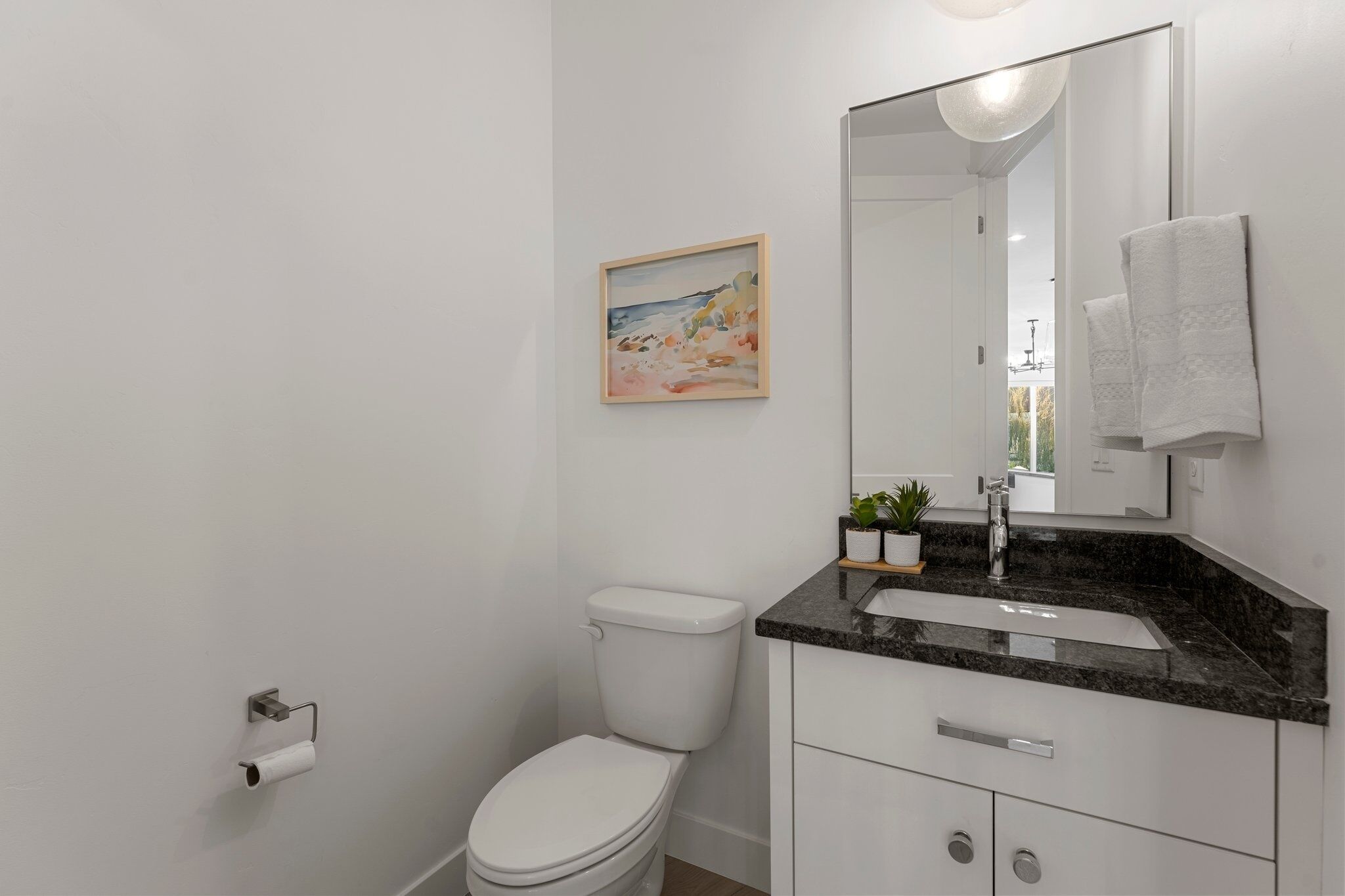Un bany modern amb un vàter blanc, un lavabo amb taulell de granit, un mirall i un quadre amb marc a la paret.