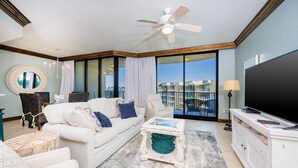 Uma sala de estar ampla com um sofá branco, uma mesa de centro e uma TV sobre um móvel branco. Há uma janela grande com vista para edifícios.