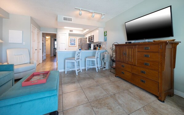 A modern living room with a large flat-screen TV, a wooden cabinet, and a kitchen area in the background.