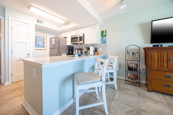 A modern kitchen with a central island, white bar stools, a wooden cabinet, and a television on the right. - Seacrest 506 1 Bedroom Condo (Gulf Shores)