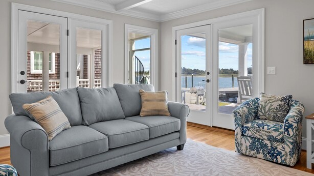 A spacious living room with a grey sofa, a patterned armchair, and a view of a balcony with a wooden deck and a body of water. - NEWLY RENOVATED Sound Front Home with Dock in Downtown Beaufort, NC (Creek Watch) (Beaufort)