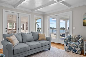 A spacious living room with a grey sofa, a patterned armchair, and a view of a balcony with a wooden deck and a body of water. - NEWLY RENOVATED Sound Front Home with Dock in Downtown Beaufort, NC (Creek Watch) (Beaufort)
