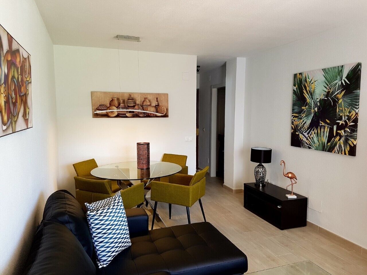 A modern living room with a glass-top dining table, yellow chairs, a black sofa, and a black console table with a lamp and a flamingo decoration.