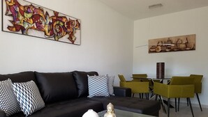 A modern living room with a black sofa, patterned cushions, a glass coffee table with decorative items, a green side table, and a dining area with green chairs and a table.
