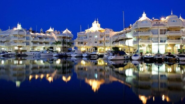 A marina with multiple yachts docked, reflecting in calm water, and a row of buildings with domed roofs and balconies.
