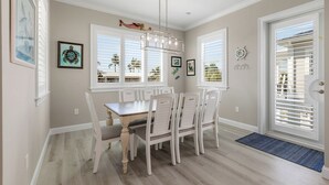 A dining area with a wooden table and white chairs, large windows with blinds, and a door with a doormat. - ARTIST HOUSE ~ Canal Front 4 BR/3.5 Ba Home w/ Pool & Ocean and Gulf Access! (Cudjoe Key)
