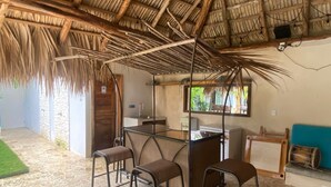 Une pièce dotée d’un toit de chaume, d’une petite kitchenette, d’une cloison en verre et de quelques chaises.